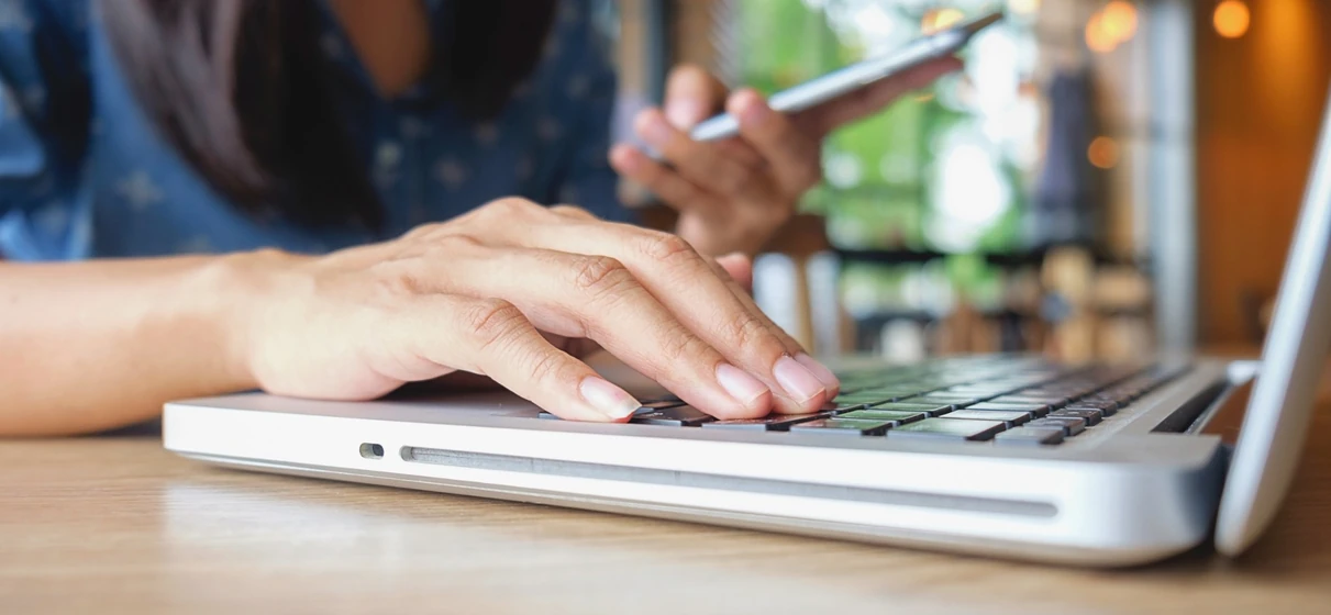 Mujer tecleando en una notebook y con un celular en la mano